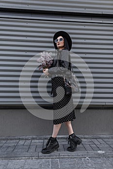 Urban woman brunette in elegant hat in sunglasses in black youth dress stands with bouquet of lilac flowers near metal vintage