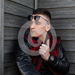 Urban young hipster man in black sunglasses in a trendy red checkered jacket with leather sleeves with a fashionable hairstyle