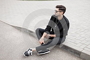 Urban young hipster man in a stylish jacket in black jeans in sneakers in trendy sunglasses is sitting on the sidewalk on a street