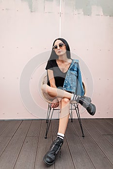 Urban young hipster woman in black leather boots in fashionable jeans clothes in stylish sunglasses posing on a straw chair