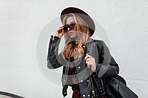Urban young hipster woman in trendy sunglasses in a chic hat in a leather jacket with a black stylish backpack