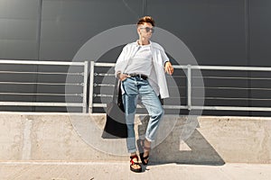 Urban young man hipster with a hairstyle in fashionable clothes with a bag stand near the metal railing near the gray building.