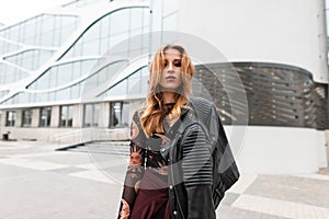 Urban young woman in a trendy patterned blouse with vintage leather jacket in pants with beautiful make-up is standing