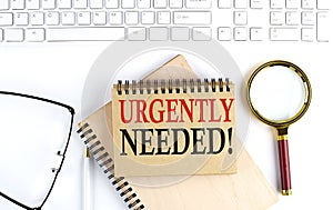 URGENTLY NEEDED text in the office notebook with keyboard, magnifier and glasses , business concept