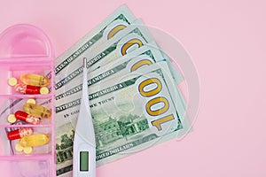 US dollar bills, an electronic medical thermometer and pills in a plastic box on a pink background