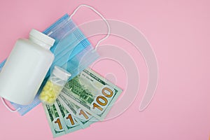 US dollar bills, packaging with pills and medicines and a medical mask on a pink background