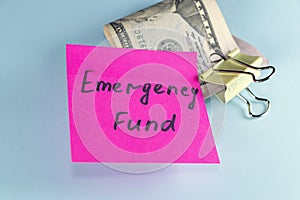 US dollars in paper clip on pastel blue background with note written EMERGENCY FUND