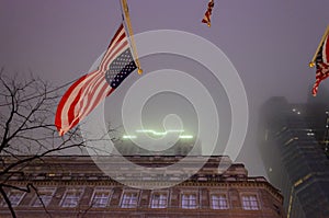 USA Flag Waving in the Wind with a Low Angle View of a Skyscraper in Background. New York City Manhattan on a Foggy Night. USA