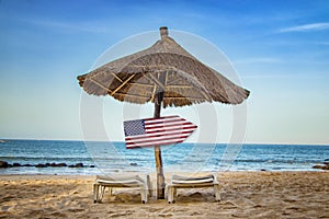 USA flag on wooden arrow sign. There are two sun loungers and a sun umbrella on the beach. It is a tropical paradise with a clear