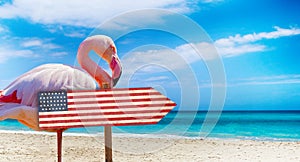 USA flag on wooden table sign on beach background with pink flamingo. There is beach and clear water of sea and blue sky in the