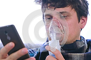 Use nebulizer and inhaler for the treatment. Sick man inhaling through inhaler mask and looking at mobile phone sitting