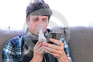 Use nebulizer and inhaler for the treatment. Sick man inhaling through inhaler mask and looking at mobile phone sitting