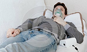 Use nebulizer and inhaler for the treatment. Young man inhaling through inhaler mask lying on the couch. Front view