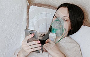 Use nebulizer and inhaler for the treatment. Young woman inhaling through inhaler mask lying on the couch and chatting photo