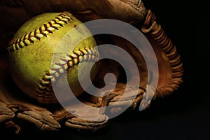 A yellow softball in an old, brown, leather glove.