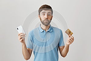 Useless credit card with low balance. Portrait of gloomy displeased young bearded guy holding smartphone and bank card