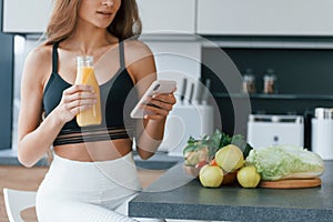 Uses smartphone. Young european woman is indoors at kitchen indoors with healthy food