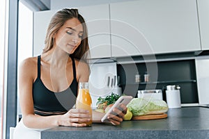 Uses smartphone. Young european woman is indoors at kitchen indoors with healthy food