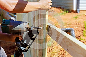 Using an air nail gun, a man constructs portions of wooden fence around his yard
