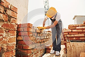 Using bricks. Young construction worker in uniform is busy at the unfinished building