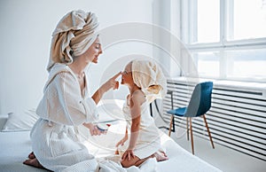 Using cream to clear skin. Young mother with her daugher have beauty day indoors in white room