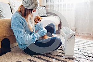 Using heater at home in winter. Woman playing with cat by electric device wearing sweater and hat. Heating