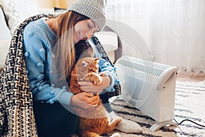 Using heater at home in winter. Woman playing with cat by electric device wearing sweater and hat under blanket. Heating