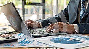 Using his laptop computer and a graph diagram, an accountant works on an office table while reading a business document