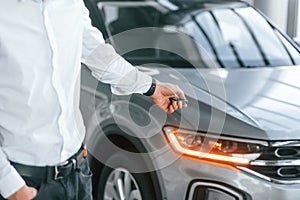 Using the keys remote controller. Young man in white clothes is in the car dealership