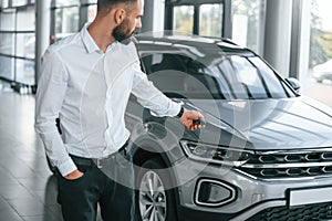 Using the keys remote controller. Young man in white clothes is in the car dealership