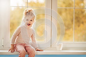 Ð¡ute little kid girl in pajama sitting by big window playing smiling enjoying home. Cacao cup standing on window. Autumn Season