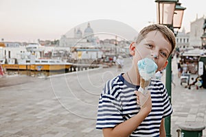 Ð¡ute seven-year-old boy in a striped t-shirt sits on the waterfront in Venice in the evening and eats gelato (ice cream