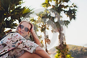 Vacation concept. Enjoying the summer. Young pretty woman in glasses sits on a palm tree over the ocean water.