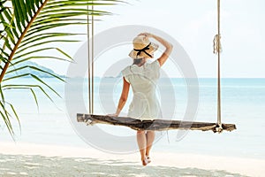 Young woman in white dress and hat swinging at a beach