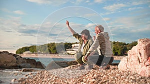 vacation for pensioners, elderly charming man and woman have fun throwing pebbles into sea while sitting on shore during