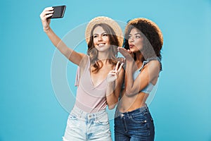 Vacation photo of two multiethnic girls wearing straw hats smiling and showing peace sign at camera, while taking selfie on smart