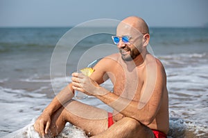 Vacationer in sunglasses holding drink decorated with pineapple and smiling sitting in wave on shore on sunny day.
