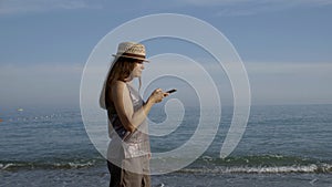 Vacationer woman with straw hat on head is walking near sea and looking at screen of smartphone photo