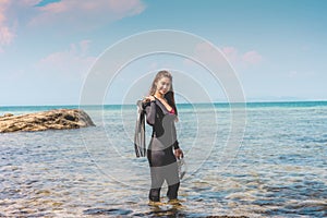 Vacations of Attractive Asian woman wearing wetsuit and holding Diving equipment fins and mask standing on seashore
