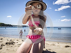 Vacations by the sea: a little girl in sunglasses and a hat pours her hands on the sand on a sea beach near the water.