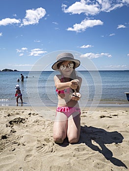 Vacations by the sea: a little girl in sunglasses and a hat pours her hands on the sand on a sea beach near the water.