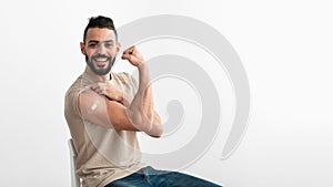 Vaccinated Arab man showing biceps muscle, demonstrating bandage after coronavirus vaccine injection on white background