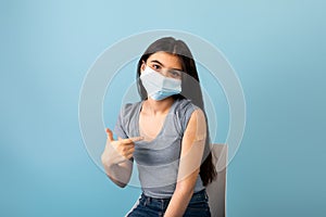 Vaccinated Indian teen girl in medical mask pointing at plaster bandage on her shoulder after getting covid-19 vaccine