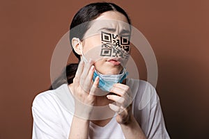 Vaccination. Portrait of a woman with a QR code instead of eyes and nose, examining her new face. Brown background. The