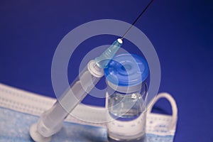 Vaccine bottle phial with no label on blue medical mask and medical syringe with injection needle. isolated on blue background.