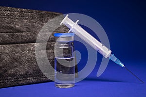 Vaccine bottle phial with no label near black medical mask and medical syringe with injection needle. isolated on blue background