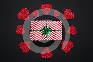 Valentine card with striped white gift box with green bow and shiny red hearts on black paper background, pattern. Symbol of