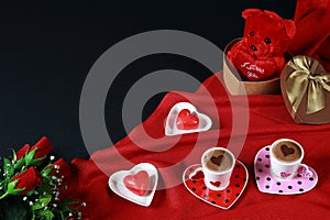 Valentine`s day concept. Coffee in heart shaped cup with candle and teddy bear in gift box with red roses on black background.