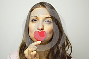 Valentine`s Day concept. Portrait of a beautiful girl kissing her small paper heart on gray background