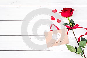 Valentine`s day congratulation. Red rose and greeting card with heart sign on white wooden background top view copy